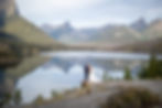 Bride and groom stand on a rocky ledge overlooking a serene mountain lake. Clear sky, reflective water, and lush green forests in the background.