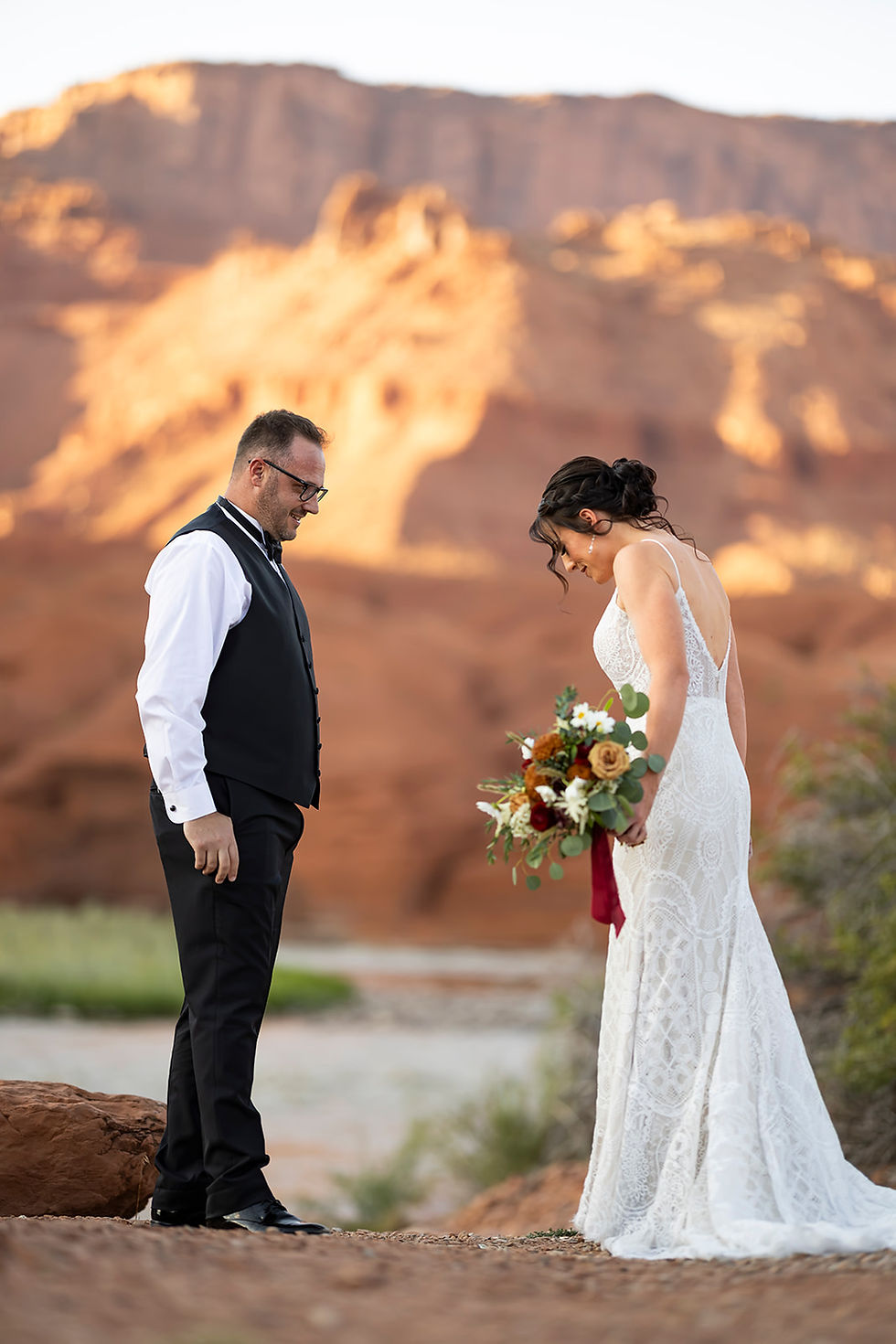 A bride in white lace holds flowers, facing a man in a vest and glasses, with a red rock desert backdrop under a clear sky. Romantic mood.