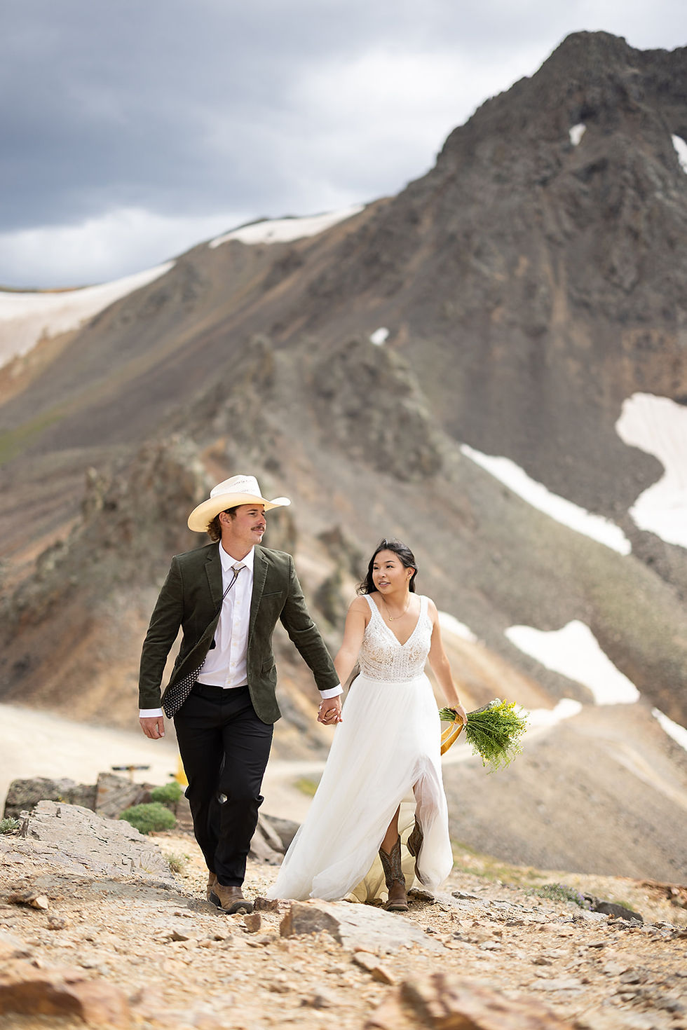 Bride in white dress and groom in cowboy hat walk hand in hand on rocky mountain path. Snow and clouds in the background.
