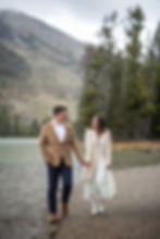 Couple holding hands, walking by a lake in the rain. Dressed warmly, surrounded by trees and mountain backdrop. Cozy and romantic mood.