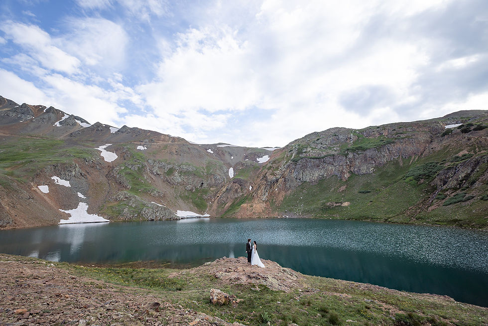 Bride and groom stand by a serene mountain lake surrounded by rocky cliffs and patches of snow under a partly cloudy sky. Romantic mood.