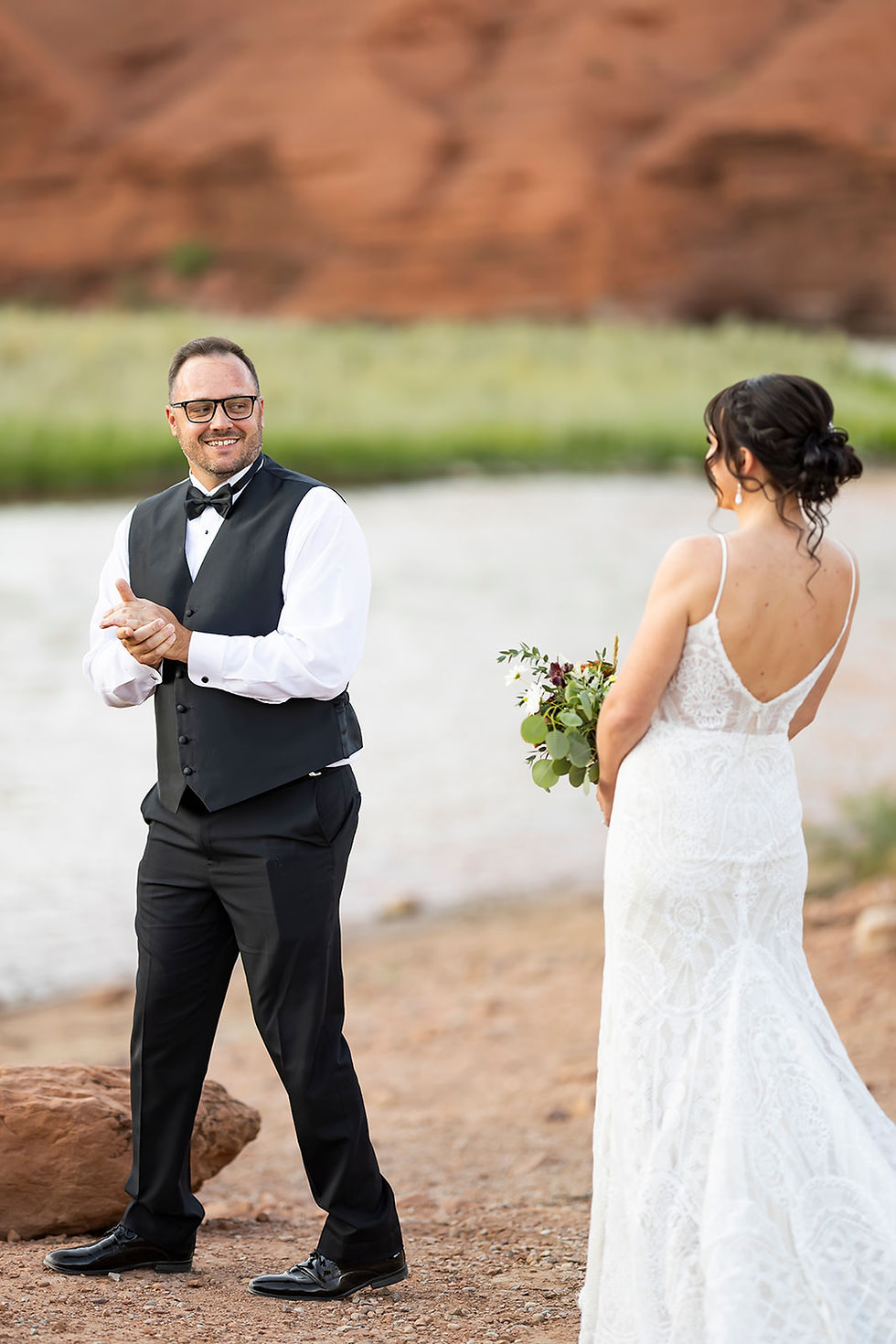 Man in formalwear claps joyfully facing a bride in lace dress holding flowers. Desert landscape with red rocks and river in background.