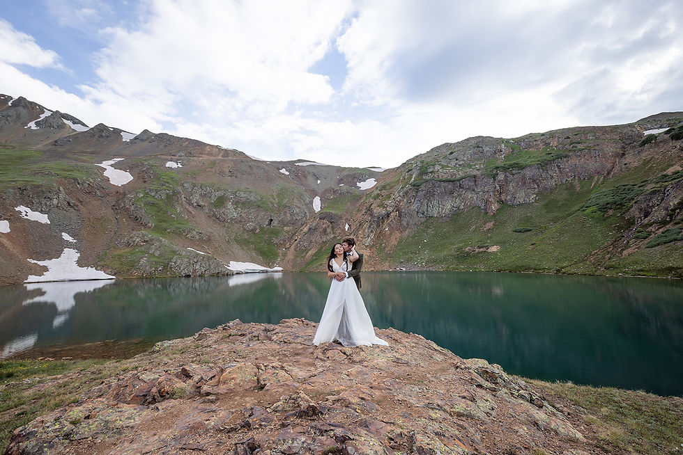Bride and groom embrace on a rocky cliff by a serene mountain lake. Snow patches on hills; cloudy sky enhances romantic, peaceful mood.