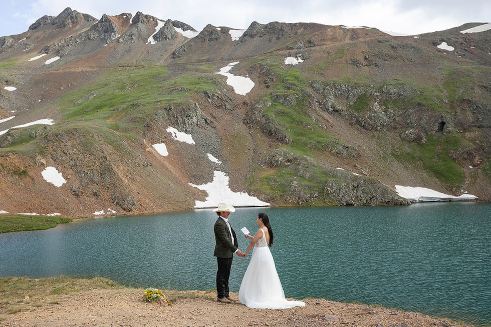 Bride and groom holding hands by a mountain lake, exchanging vows. Snow-dusted peaks in background; serene and picturesque setting.