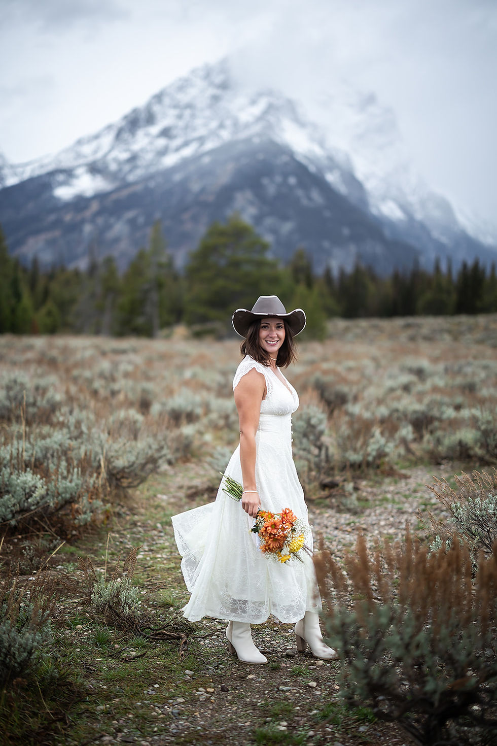 A woman in a white dress and cowboy hat smiles, holding flowers in a mountainous landscape with cloudy skies and green foliage.