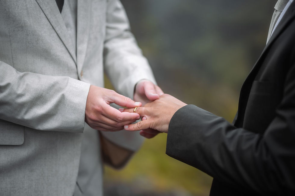 Two people in suits exchange a gold ring during a wedding ceremony, with a soft blurred background and a solemn mood.