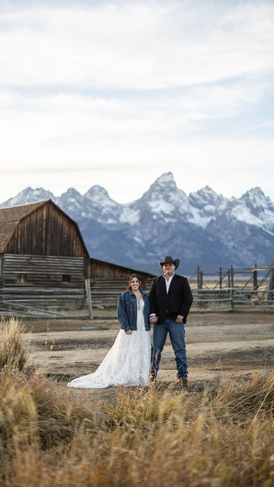 Cowboy and bride kiss in front of rustic barn, with snowy mountains in the background. Golden grass, clear sky, romantic mood.