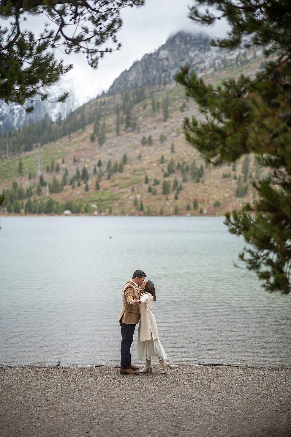 Couple embracing by a lake, surrounded by trees and mountains. They're on a pebbled shore, dressed warmly. The mood is serene and romantic.