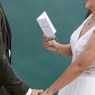 A couple holds hands at a lakeside in a mountainous setting. The man in a cowboy hat reads vows; the woman smiles in a white dress.