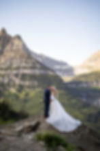 Bride and groom embrace on a rocky ledge with majestic mountains in the background, her white dress flowing, under a clear blue sky.