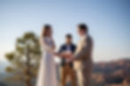 A couple exchanging vows outdoors, with a third person officiating. The bride is in a white dress and emotional. A tree and mountains in the background.