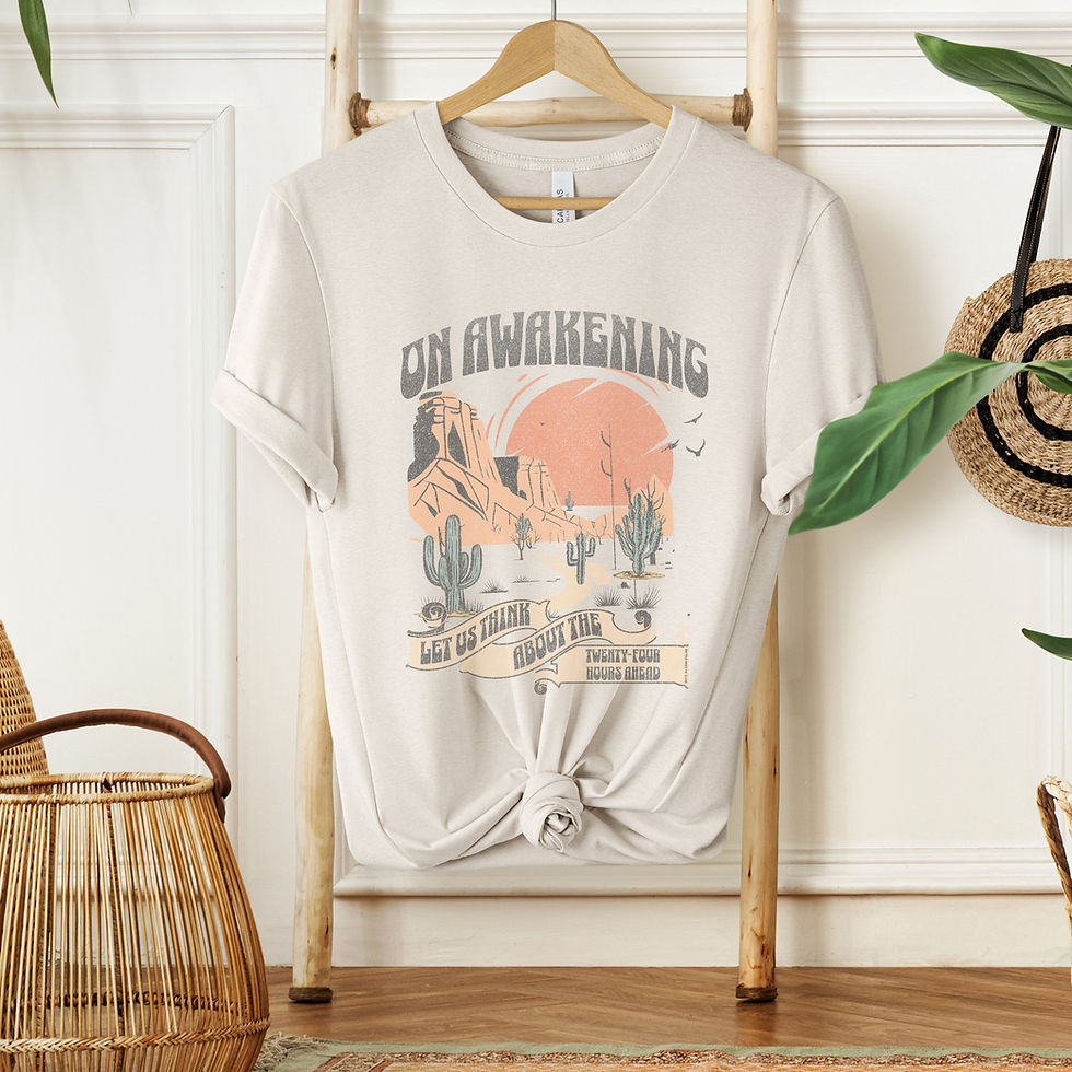 Thumbnail: Cream colored shirt with muted toned scenery with desert moutains, cacti, and orange sun with "on awakening let us think about the 24 hours ahead" on a rack with wicker baskets and plant