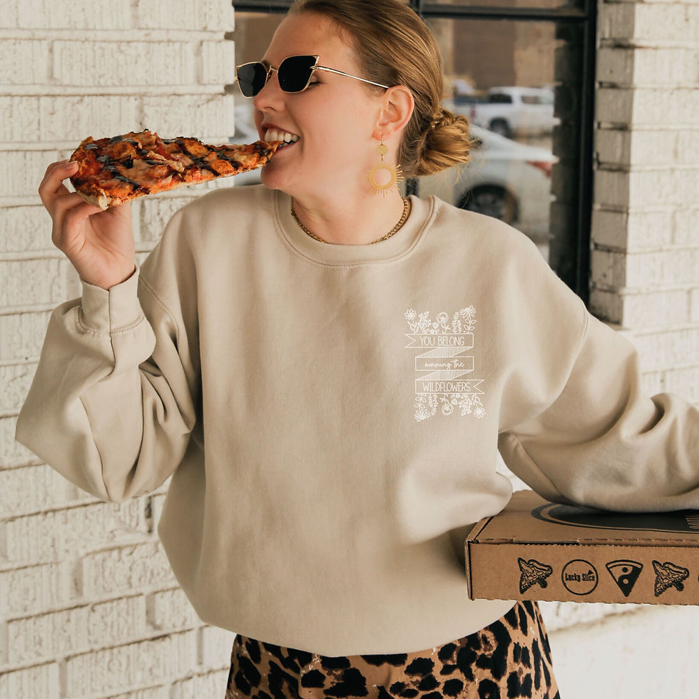 Thumbnail: Woman eating pizza wearing tan crewneck with "you belong with the wild flowers" on the front over the chest with logo on the arm "you belong"