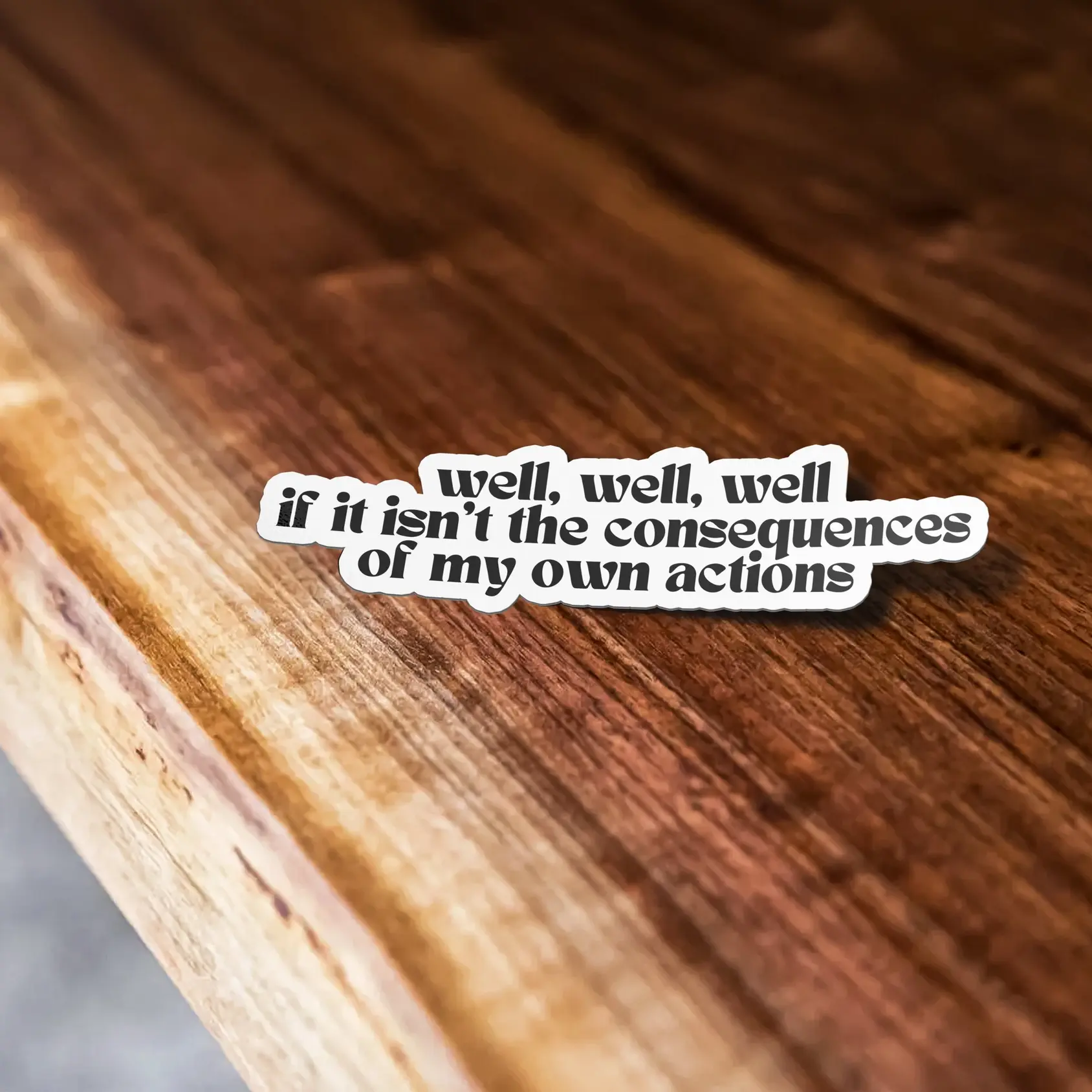White sticker with black boho writing" well, well, well, if it isnt the consequences of my own actions" on a wood table.