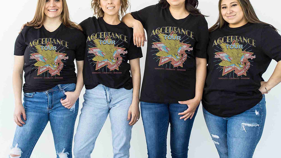 Four women in black "Acceptance Tour" T-shirts pose together, smiling. They wear blue jeans and stand against a plain white wall.
