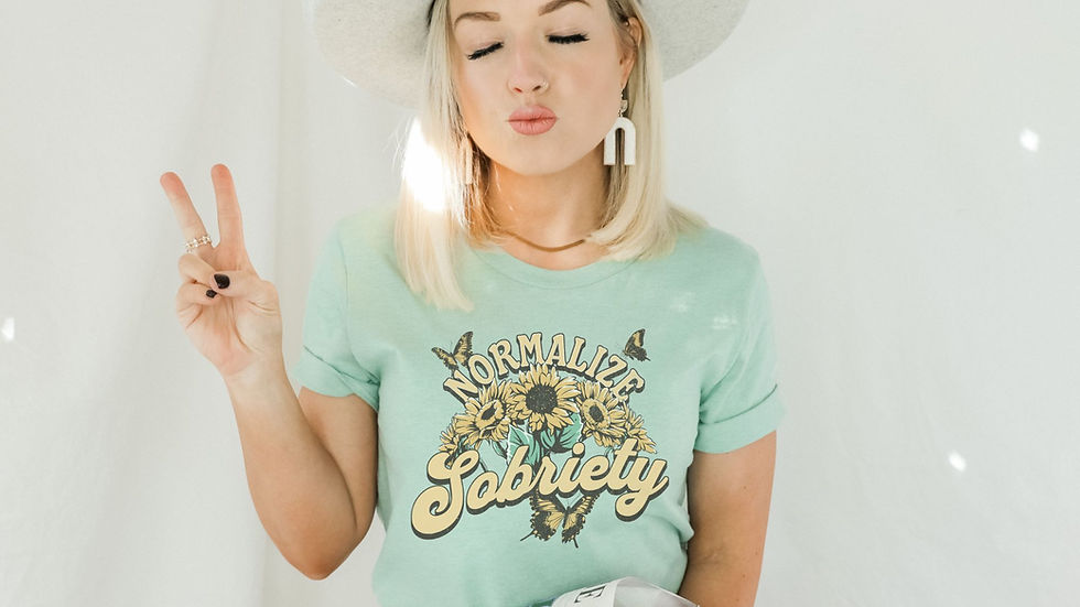 Woman in a light green "Normalize Sobriety" shirt and gray hat makes a peace sign, eyes closed, holding a colorful bouquet, on a white background.
