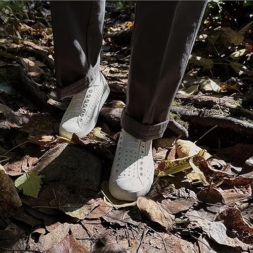 Person wearing white Native shoes walking on a rocky forest floor with fallen leaves.