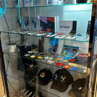 Glass display cabinet filled with hardware crypto wallets, bitcoin badges, cups, hats and tops.