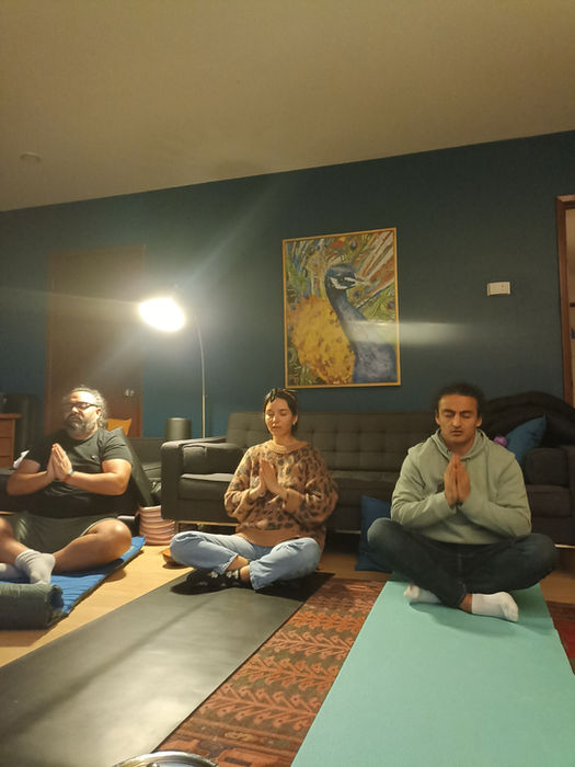 Three people meditating indoors on mats; a peacock painting on the wall.
