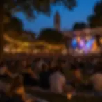 Outdoor evening concert with a seated crowd on grass, colorful stage lights, and a church clock tower under blue twilight.