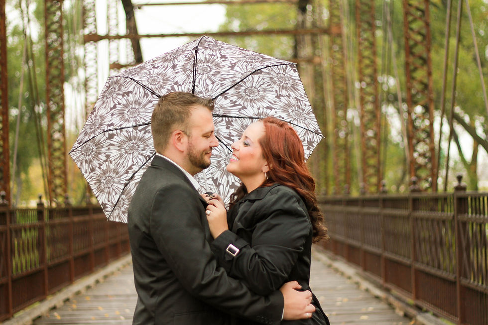 A couple looks at each other under an umbrella on a wooden bridge with greenery behind them.