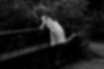 A curly haired barefoot woman in a white night dress on a bridge looks over the fence of the bridge down toward the water - black and white