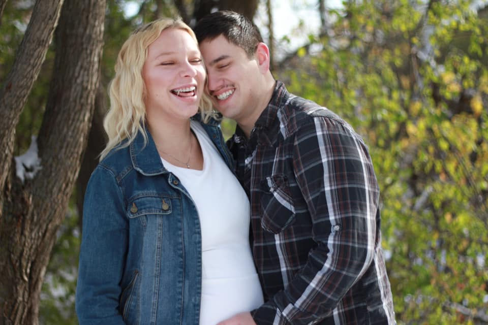 A blonde woman and a brunette man playfully laugh as he hugs her