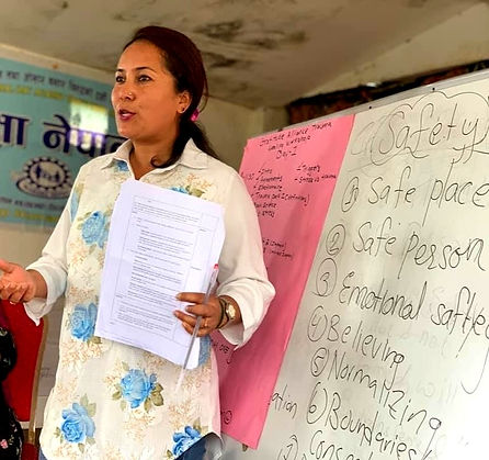 Nepali woman facilitator standing in front of a white board teaching about Safety, with Safety written behind her on the board, along with "safe place, safe person, emotional safety, believing, normalizing..."