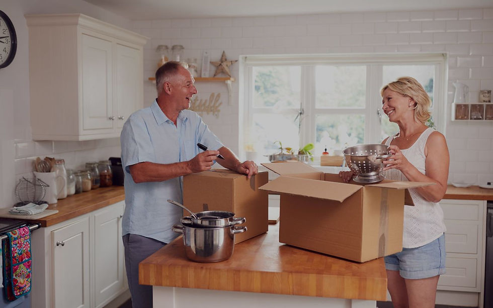 A man and a woman smiling while packing kitchen items in cardboard boxes on a wooden countertop. sell your home for cash buffalo