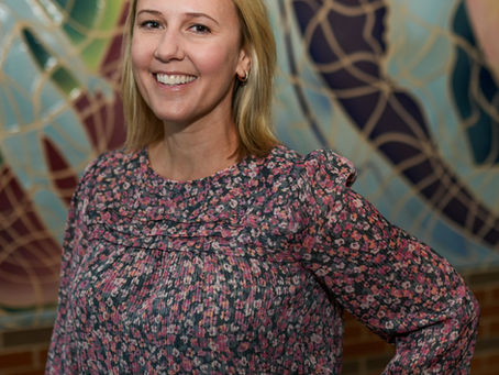 A white woman with a blonde bob and floral blouse smile in front of a multi-colored tile mural.