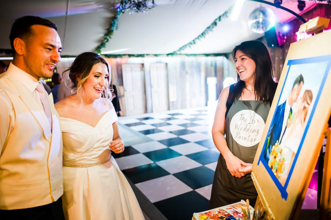 A bride and groom smile happily whilst an artist shows them their painting