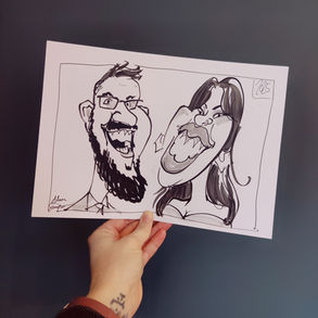 A hand holds up an image against a dark green background. The image is a caricature of a man and woman drawn in black marker pen.