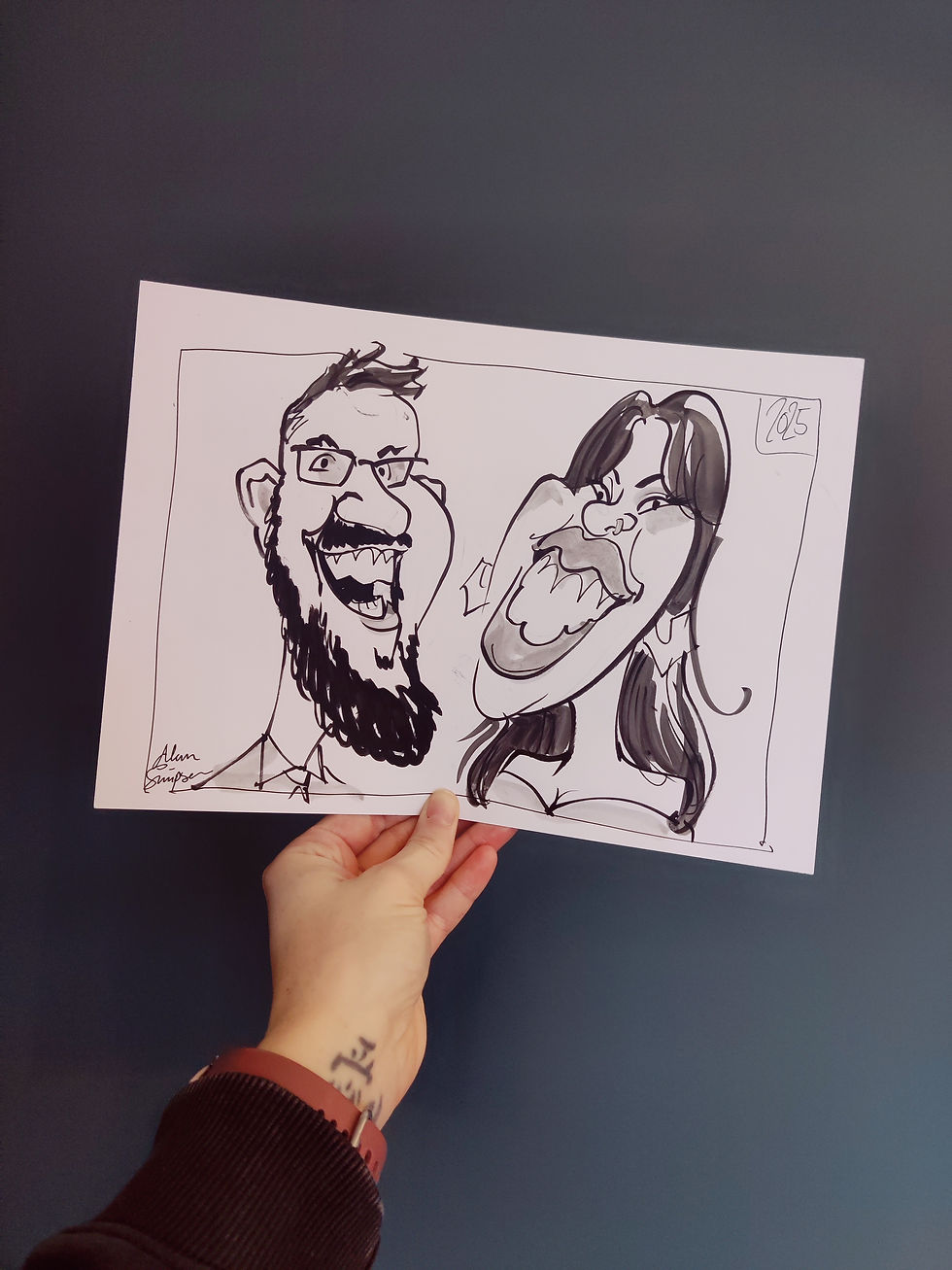 A hand holds up an image against a dark green background. The image is a caricature of a man and woman drawn in black marker pen.