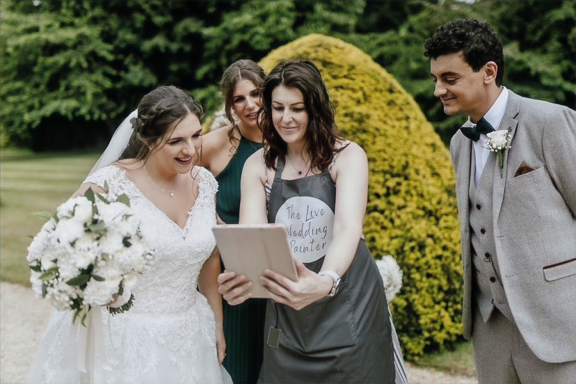 A bride, groom and bridesmaid gather around an artist who is showing them images on a tablet.