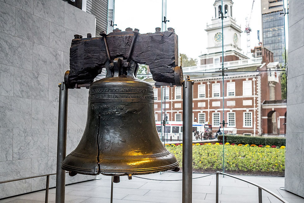 The Liberty Bell with a crack, displayed indoors. Background shows a historic brick building with a clock tower, viewed through glass.