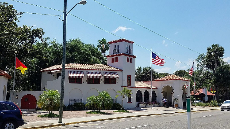 Spanish-style building with red roof tiles, striped awnings, and American flags. Sunny day with palm trees and a clear blue sky.