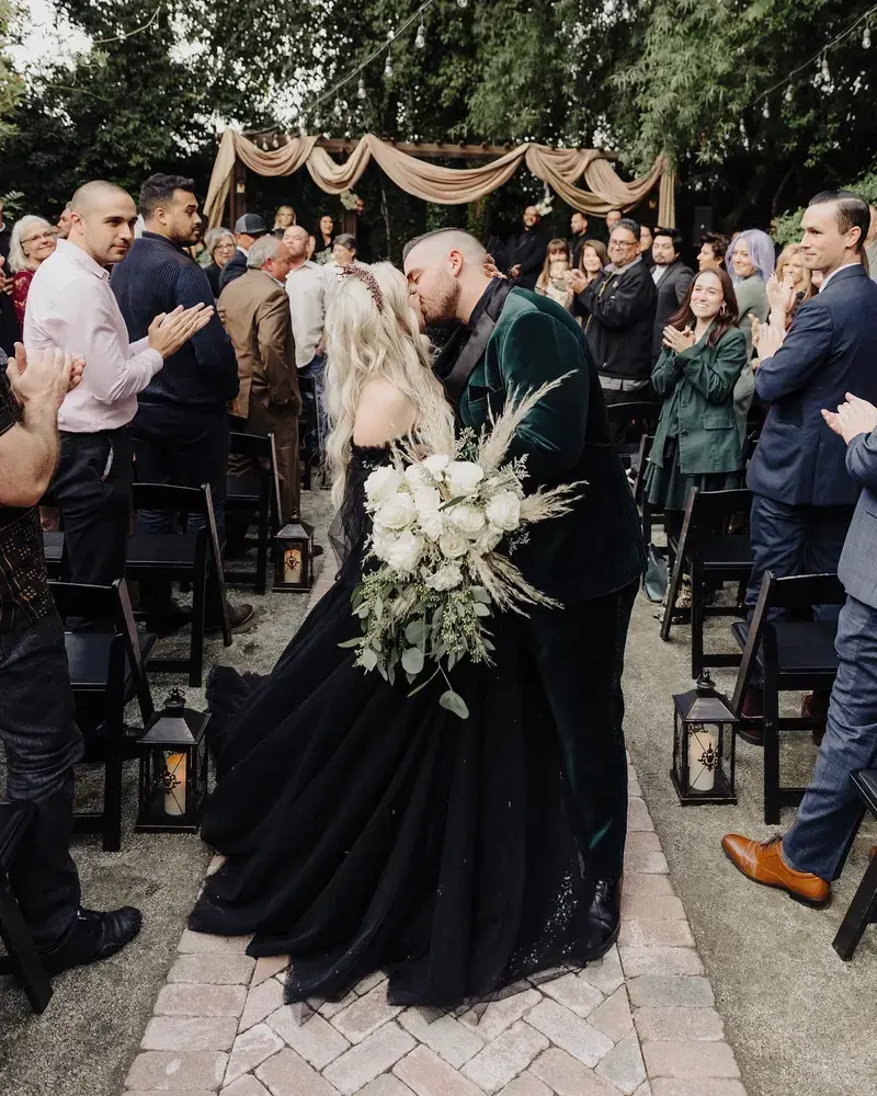 A couple shares a romantic kiss at their wedding ceremony, with the bride in a stunning black gown and the groom in a velvet suit. Guests stand and applaud warmly against a backdrop of elegant drapery and greenery.