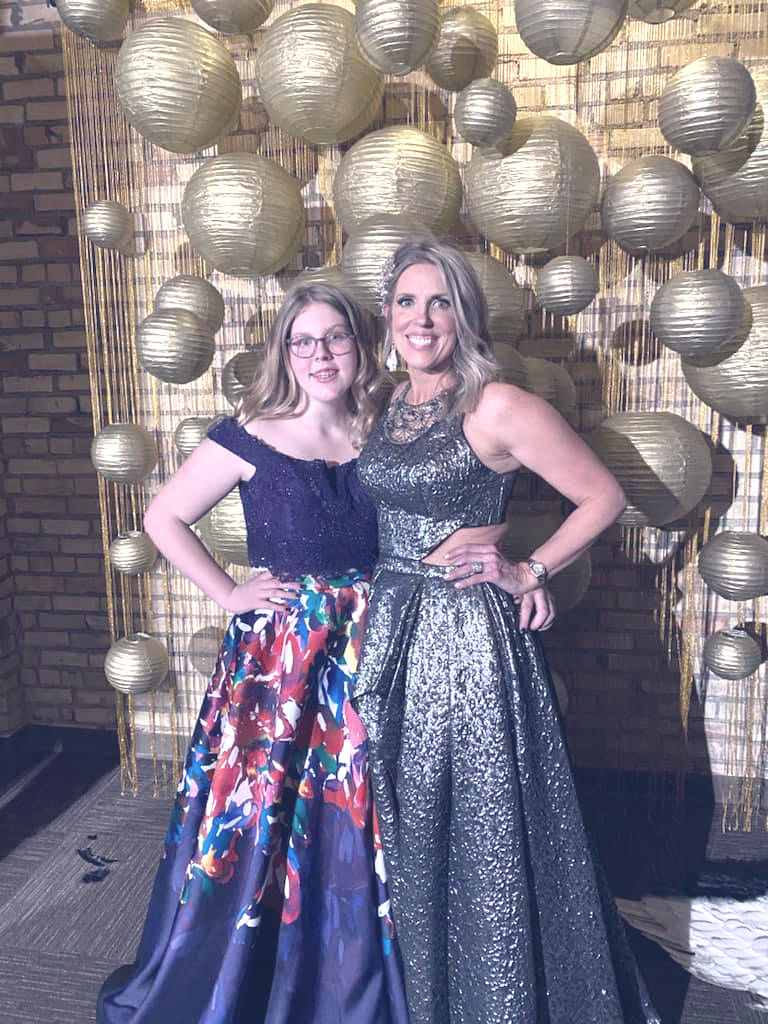 Two women in elegant gowns pose smiling in front of a backdrop with gold spherical decorations. The setting is festive and celebratory. Melanie Rudd
