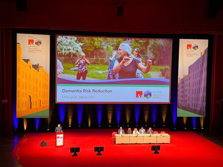 A person standing at a podium speaking to a large conference room. Banners and a large projector screen behind them.