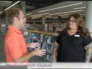 Micala Gingrich-Gaylord speaking with Avery Osen from Good Day Kansas at a Wichita Public Library among the shelves of books.