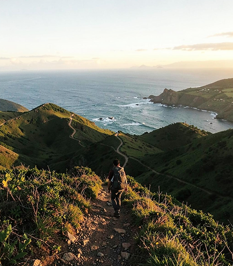 A hiker on a winding coastal ridge trail with rolling green hills and the sea below.png