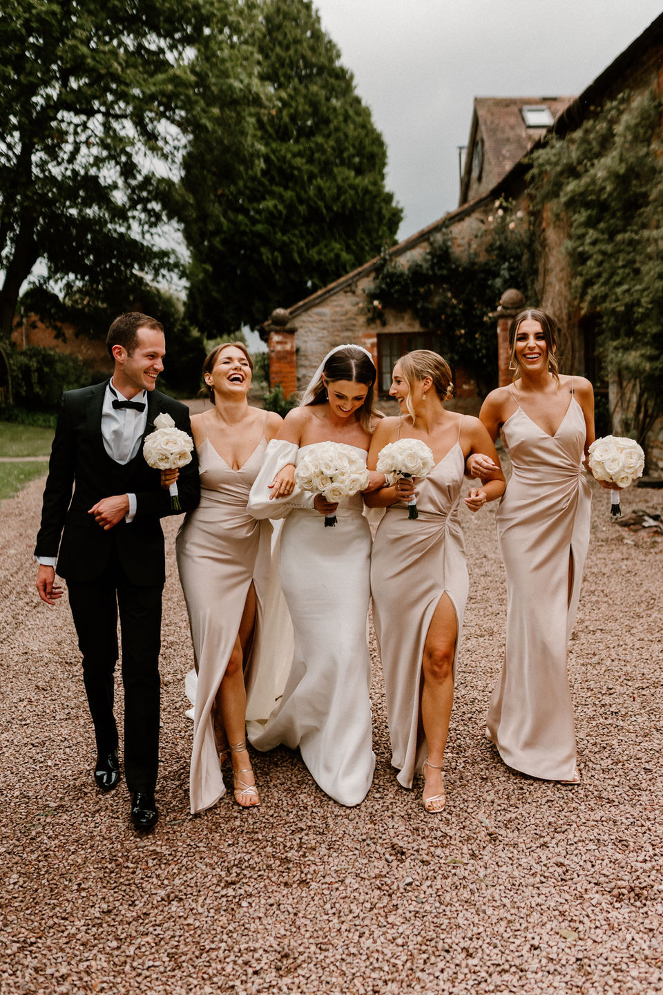 This vibrant image shows a joyful bride walking alongside her bridal party at Birtsmorton Court. The bride is dressed in a sleek, elegant white gown with off-the-shoulder sleeves and carries a bouquet of white flowers. She is surrounded by her bridesmaids, who are wearing champagne-colored dresses with thigh-high slits, each carrying matching bouquets.
The group walks arm-in-arm, laughing and radiating happiness. A groomsman, dressed in a sharp black tuxedo with a bow tie, walks beside them, holding a bouquet as well. The setting features a rustic gravel pathway with lush greenery and the charming architecture of Birtsmorton Court in the background. The scene feels warm and celebratory, capturing a candid and fun moment of friendship and joy.