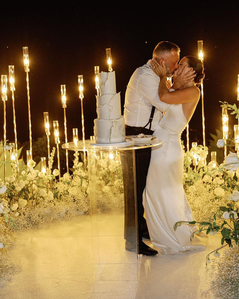 Bride and groom kissing beside their wedding cake, surrounded by flickering candlelight and lush white floral decor.