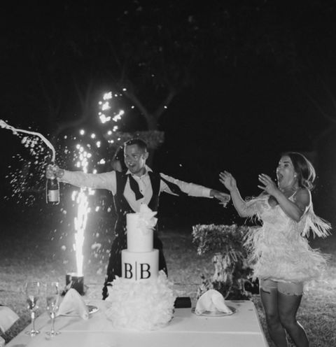 Bride and groom celebrating outdoors at night with champagne and sparklers beside their wedding cake.