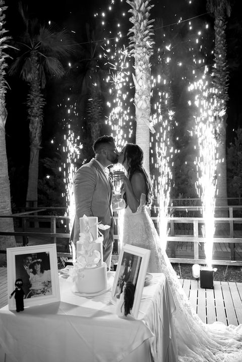 Black and white photo of a couple cutting their wedding cake under palm trees and sparkling night-time fireworks.