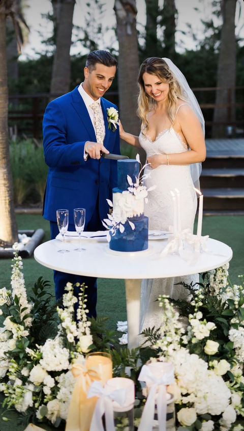 Bride and groom cutting their blue wedding cake together at a round outdoor table on a sunny afternoon.