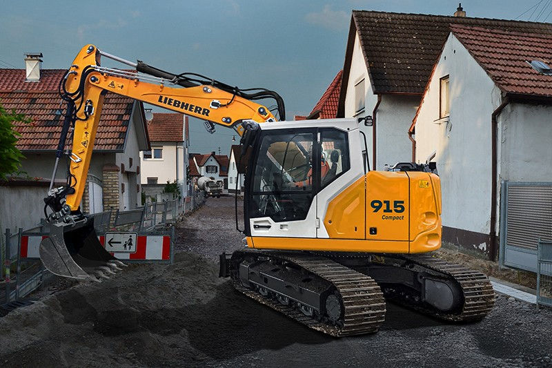 Yellow Liebherr 915 Compact excavator on a narrow residential street beside houses, digging dark soil near a road sign.