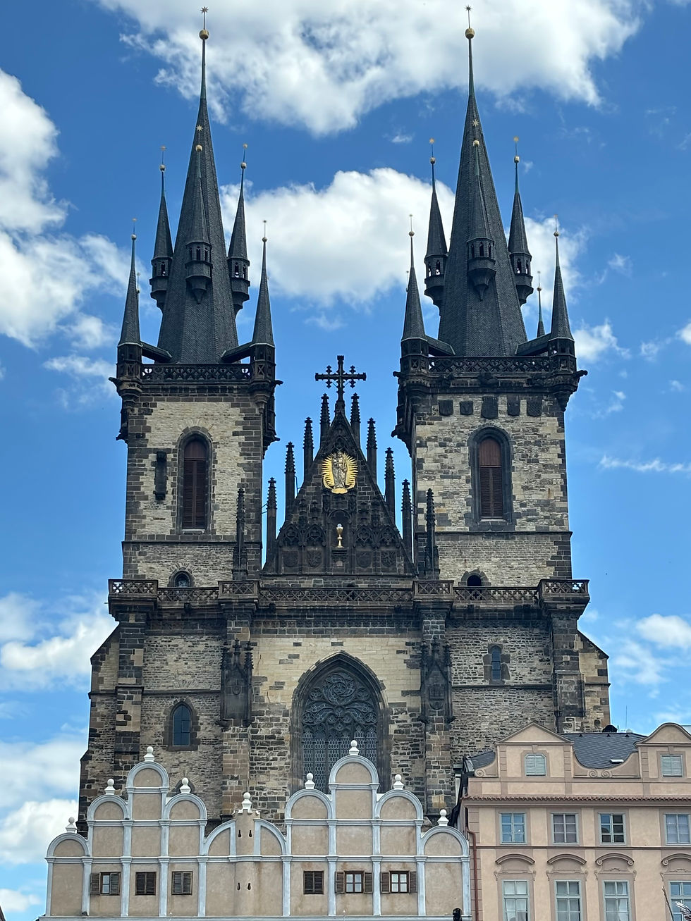 Gothic church with twin spires and stone facade against a bright blue sky with clouds, framed by pastel buildings below.
