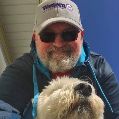 Selfie of a white man with sunglasses, a ball cap and hoodie smiling down at the camera with a small white fluffy dog in the frame.