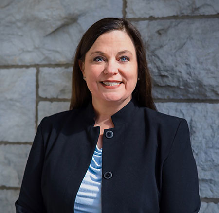 Headshot of a smiling white woman with brown hair in a blue blazer against a background of bricks.